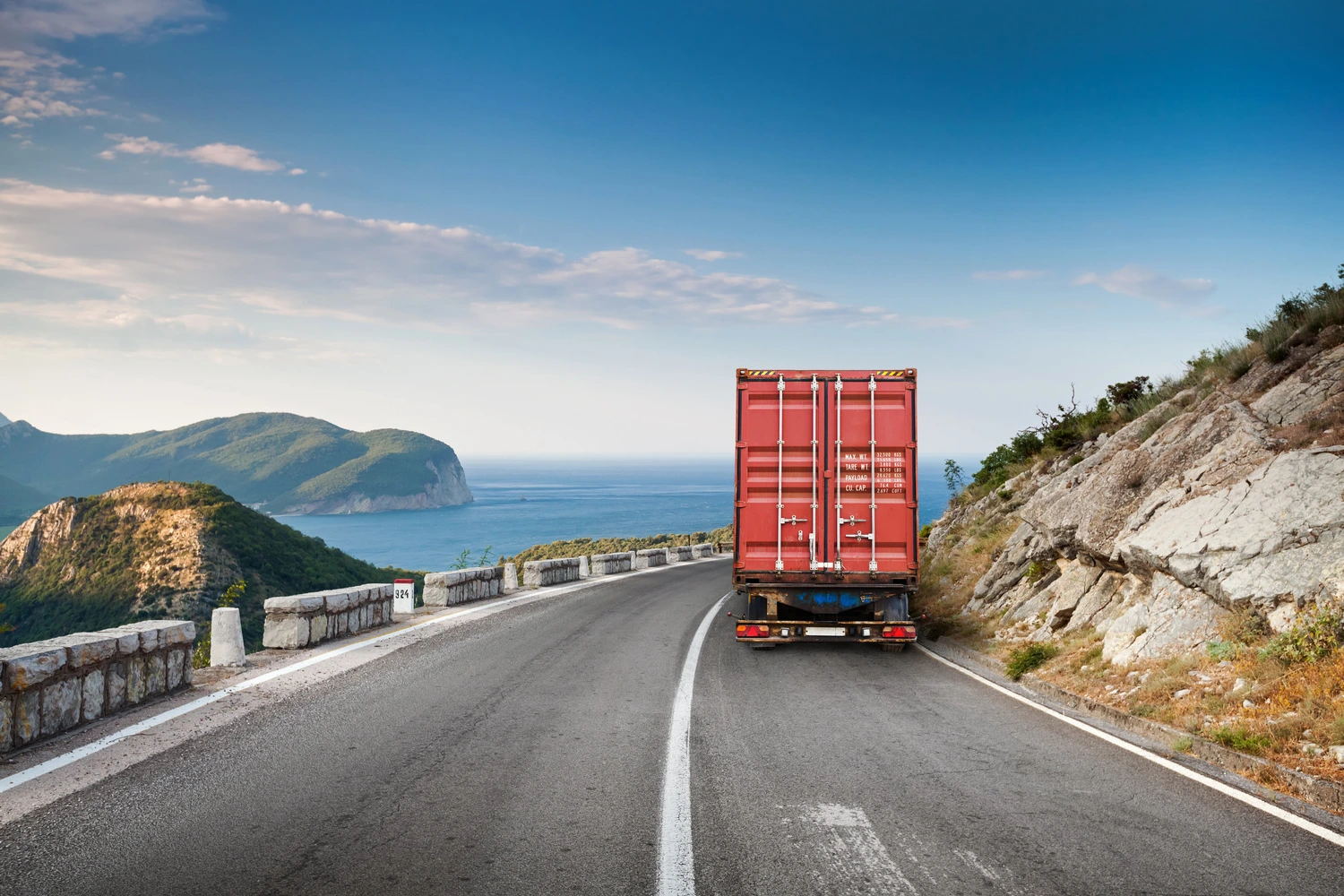 Cargo truck driving along a coastal mountain road with cliffs and ocean in the distance